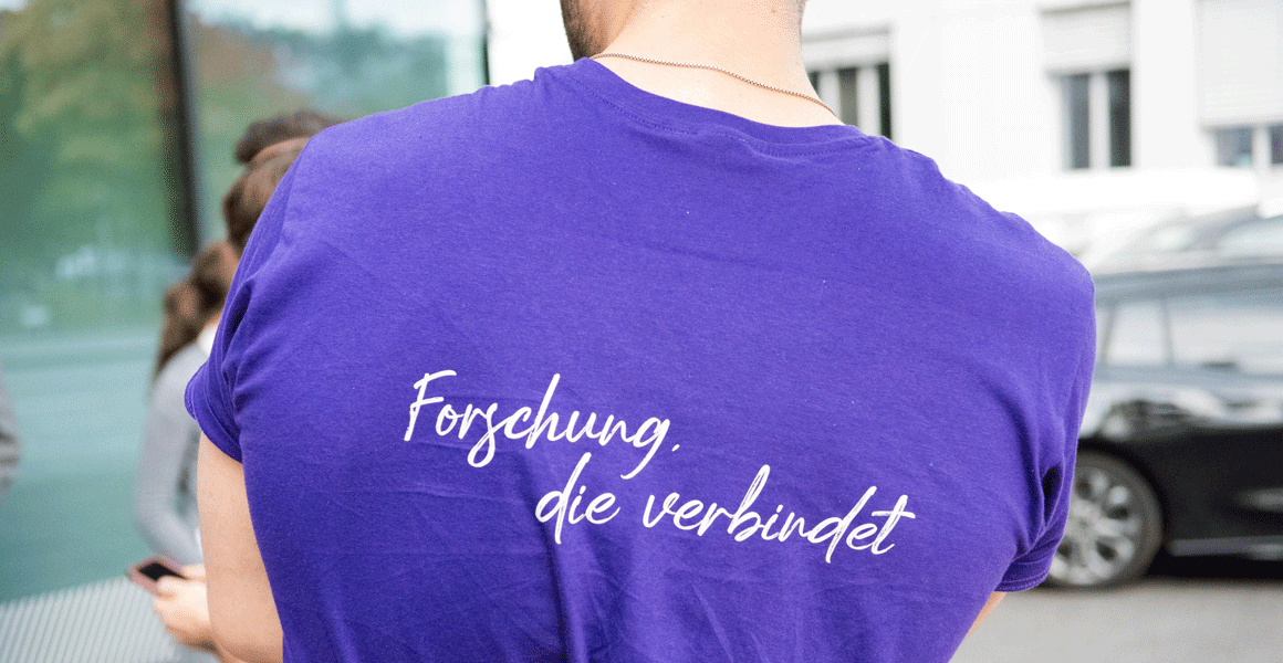 Rückansicht: Mann mit lila T-Shirt mit COREMED-Slogan "Forschung, die verbindet"