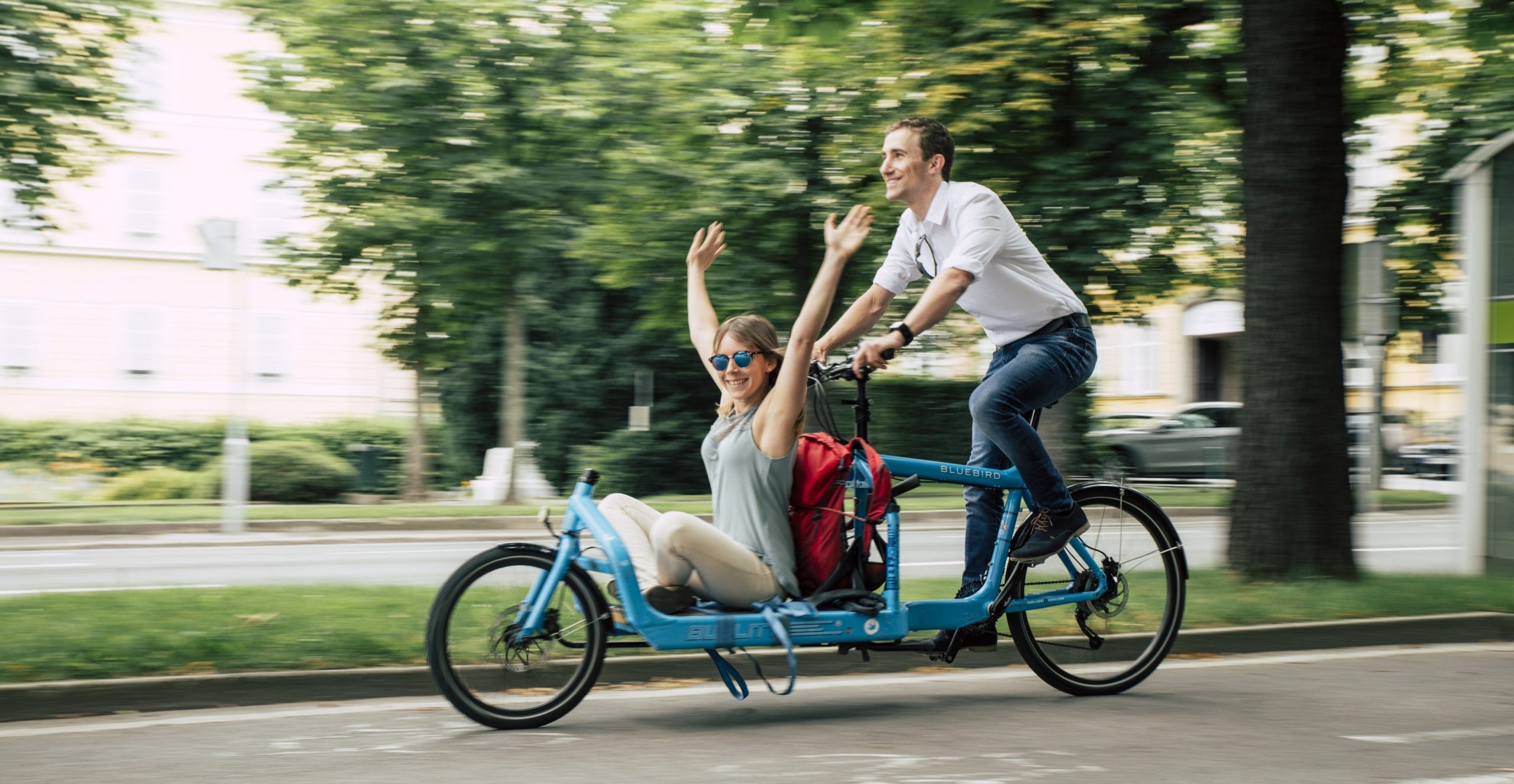 Mann transportiert Frau auf einem blauen Lastenrad, welche die Arme nach oben streckt und lächelt.