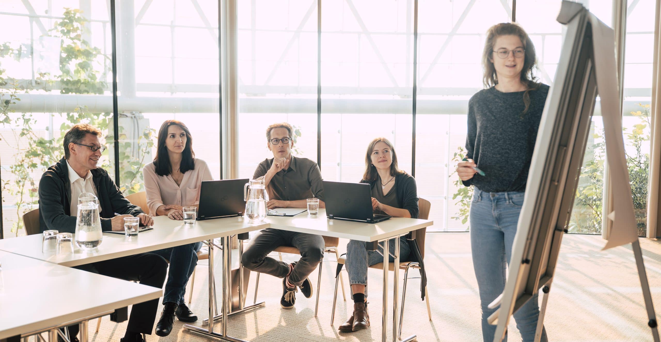 Frau präsentiert auf einem Flipchart, dahinter sitzen zwei Frauen und zwei Männer, die aufmerksam zuhören. Auf dem Tisch sind zwei Laptops und Wasserkrüge mit Gläsern. Symbolbild für Nachhaltigkeitsbewertung