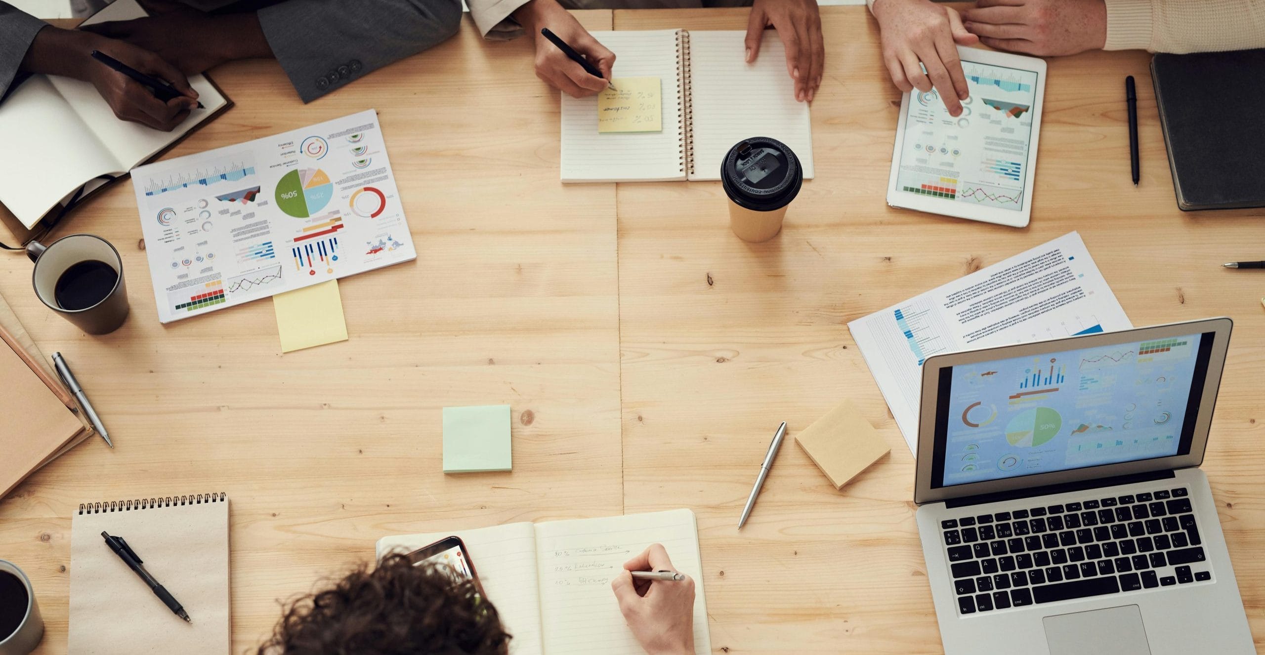 Blick von oben auf einen Tisch, rundherum sitzen Menschen und nehmen an einem Meeting teil. Ein Laptop, ein Tablet und div. Schreibunterlagen sind am Tisch.