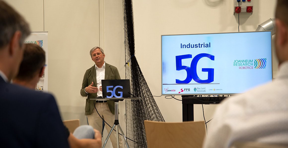 Moderator vor dem Monitor, der ein 5G Symbol zeigt