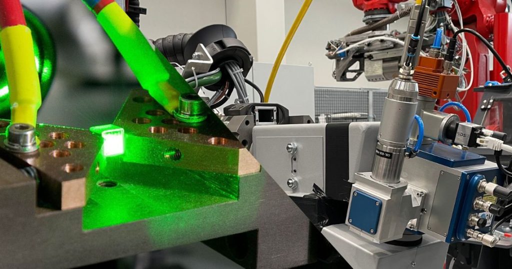 Ein grüner Laser beleuchtet einen präzise eingespannten Kristall in einem optischen Laboraufbau und ein robotergeführter NDT-Testkopf