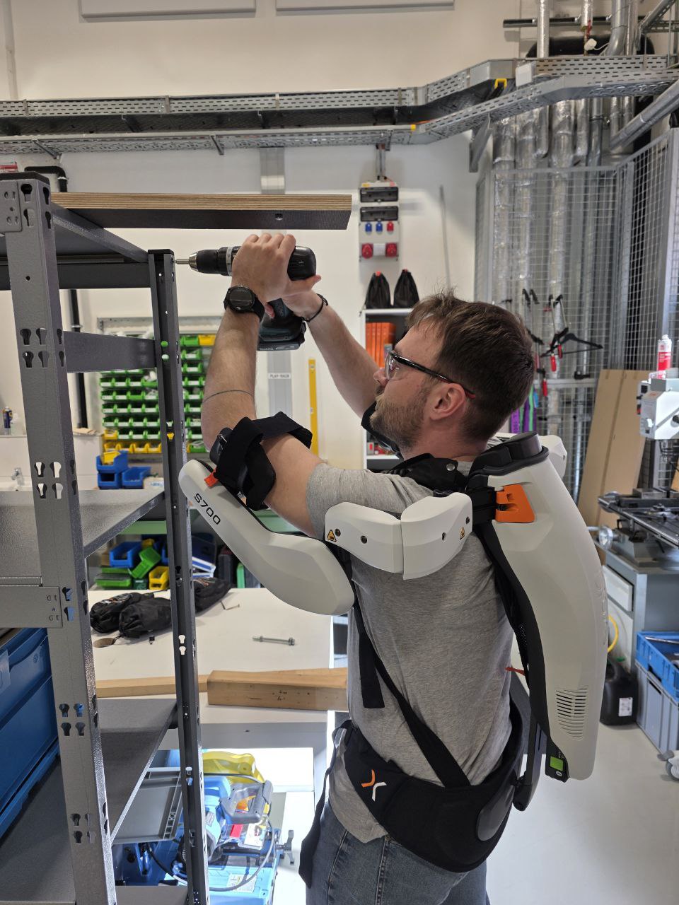 Ein Mitarbeiter steht vor einem Regal und trägt ein Exoskelett für den Oberkörper und hält einen Akkuschrauber an das Regal