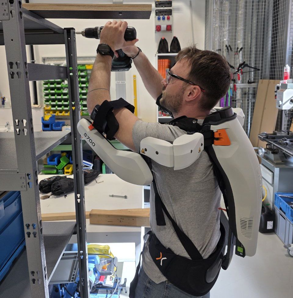 Mitarbeiter steht vor Regal und trägt ein Exoskelett für den Oberkörper